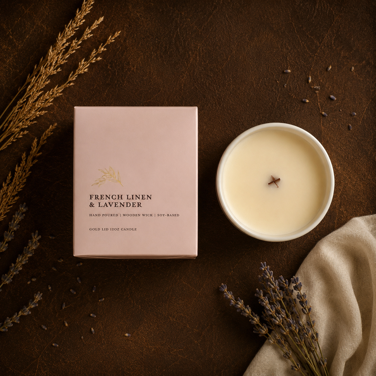 A wax candle shown in a white round vessel next to its blush pink box labeled 'FRENCH LINEN & LAVENDER,' arranged on a dark brown textured surface with dried lavender and beige fabric props.