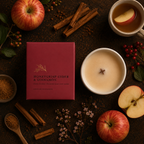 A boxed candle labeled 'HONEYCRISP GIDER & CINNAMON' sits on a dark rustic surface surrounded by apples, cinnamon sticks, a mug of tea, and a lit candle in a white bowl, creating a warm autumnal scene.