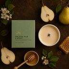 A styled flat-lay of a candle product with its sage green box and a white candle bowl surrounded by pear halves, honeycomb, honey dipper, honey, white blossoms, and green leaves on a dark brown textured surface. The box reads 'ORCHID PEAR & HONEY'.