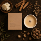 A candle product styled in a flat lay on a dark textured surface, with the kraft paper box labeled 'SANTAL & SHEA BUTTER' beside a lit white candle. Surrounding props include cinnamon sticks, a bowl of solid wax, dried flowers, nuts, and leaves.