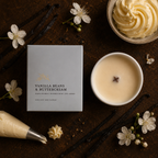 A vanilla bean and buttercream scented candle shown with its light gray box, a lit cream-colored candle in a white bowl, vanilla beans, white flowers, and a piping bag on a dark rustic surface.