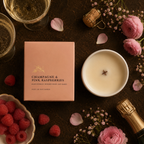 A styled flat-lay of a candle product with its blush pink box, next to a lit white candle, champagne, raspberries, pink flowers, and cork props on a dark textured surface.