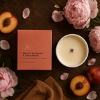 A peach-colored candle box labeled 'PEONY BLOSSOM & NECTARINE' is arranged on a dark brown surface beside a white candle vessel with creamy wax and a wick. Pink peony flowers, peach halves, and scattered petals surround the product in a styled flat-lay composition.