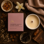 A styled flat-lay of a candle product showing a pink box labeled 'CASHMERE VANILLA & WARM AMBER' next to a white candle vessel with cream wax and a wooden wick. The scene includes vanilla beans, amber pieces, dried flowers, and a brown cloth on a dark surface.