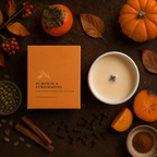 A hard wax candle in an orange box labeled 'Pumpkin & Persimmons' is arranged on a dark rustic surface with autumn props including a white candle vessel, cinnamon sticks, cloves, pumpkin, persimmon, leaves, and seeds.