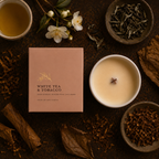 A square blush pink candle box labeled 'WHITE TEA & TOBACCO' is arranged in a styled flat lay on a dark rustic surface, surrounded by tea leaves, dried leaves, a cup of tea, white flowers, and a white candle with a single wick.